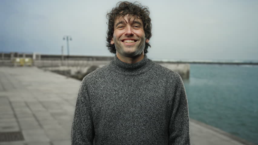 Young man making rock gesture at seaside in grey sweater, smiling in outdoor beach setting with ocean background.