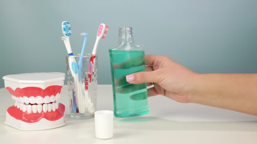 Hand pouring mouthwash into cap with toothbrushes and dental model on table