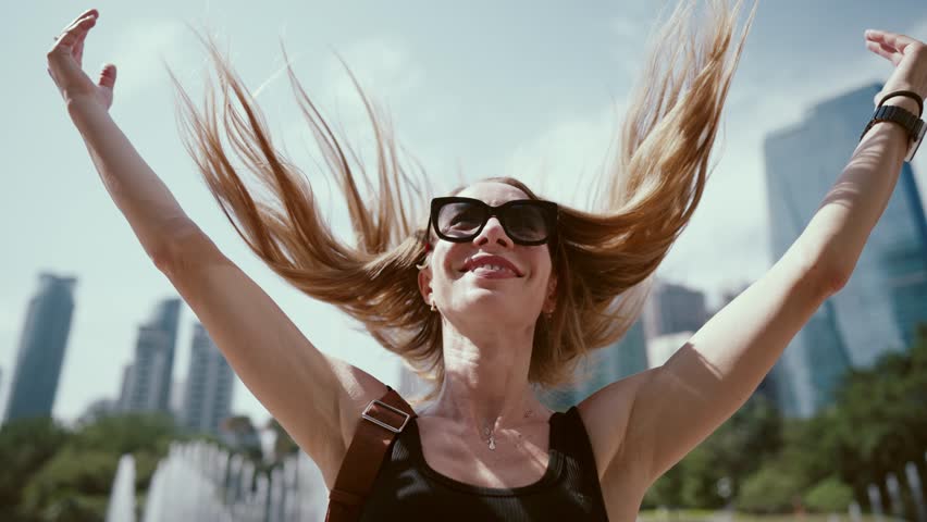 Happy woman playing with her long blonde hair in urban view in summer, portrait. Positive emotions and good mood, smiling female person enjoying life and sunny weather, be joyful and have fun forever
