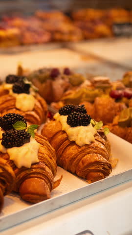 A variety of fresh, golden croissants and pastries are displayed in a bakery showcase. The croissants are topped with colorful fruits and cream, inviting customers to indulge in these delicious treats