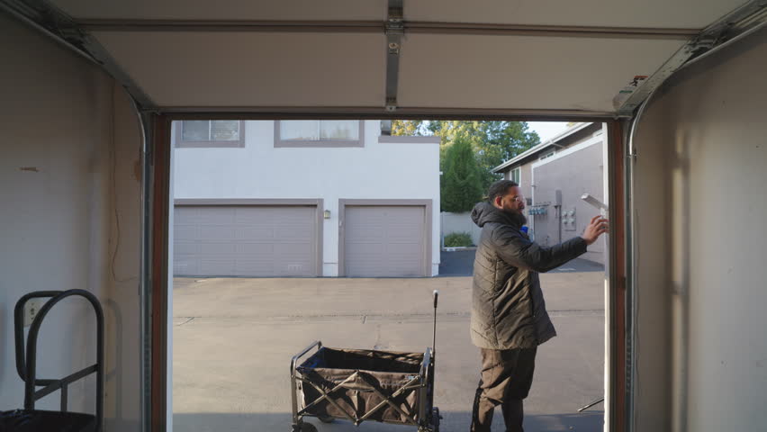 A man steps out from a garage while pulling a folding wagon behind him, showcasing the significance of routine activities and the useful applications of such items in a conventional home setting