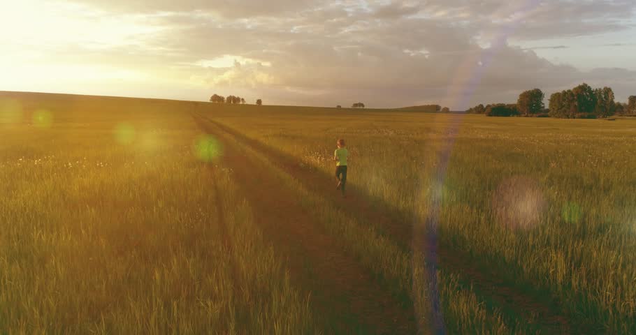 Aerial shoot of a sporty child runs through a wheat field. Evening sport training exercises at rural meadow. A happy childhood is a healthy way of life. Outdoor runing traning. Radial movement, sun