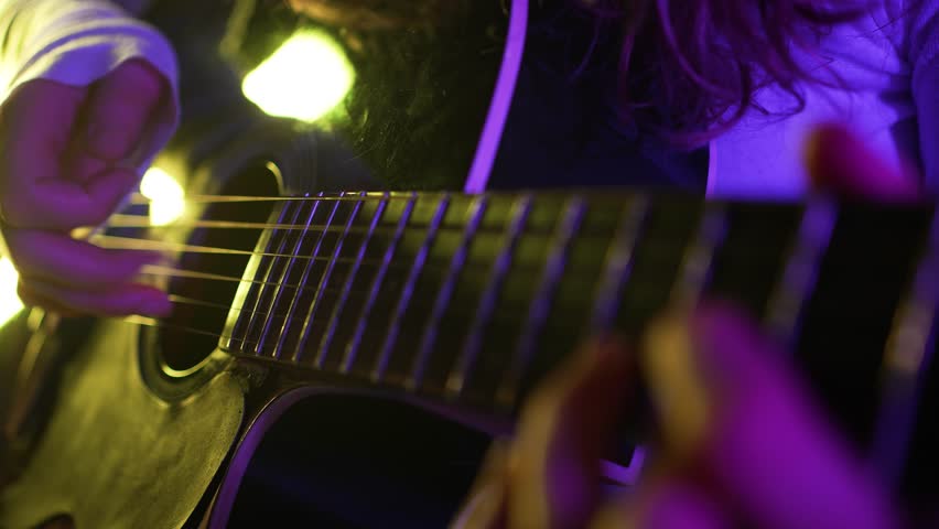 Close-up of musician's hands playing acoustic guitar, illuminating purple and yellow stage lights casting moody shadows while capturing intricate musical performance and emotional expression