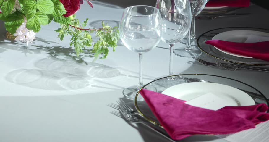 Close-up of a festive table setup with wine glasses, white plates, and pink napkins. Soft artificial lighting highlights the reflections. Handheld shot with cozy and elegant atmosphere. 4k footage