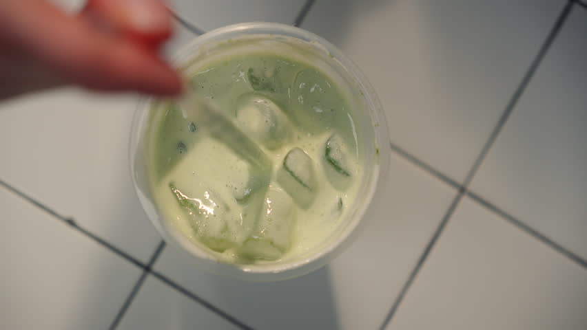 Cinematic shot top down tabletop view of iced tea or coffee with matcha, which a girl hand stirs with drinking straw.