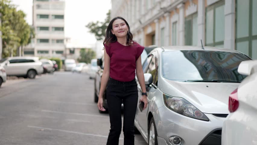young asian woman joyfully walks and jumps with excitement toward her car, smiling and raising arm in celebration, cheerful urban scene with positive energy and freedom