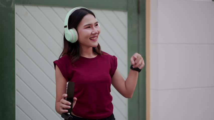 A young Asian woman in casual outfit listening to music with wireless headphones, smiling and pointing up while holding smartphone outdoors, expressing joy and rhythm in urban setting