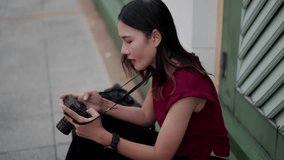 Young asian woman sitting on pavement, smiling while browsing images on digital camera screen, relaxing during urban photo walk with casual outfit and gear - Powered by Shutterstock - Get 15% off with code: PIKWIZARD15