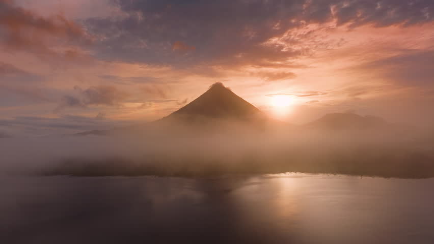 Experience stunning aerial views of the sunrise over Laguna de Arenal and Volcan Arenal in Costa Rica, surrounded by mist and greenery, creating a tranquil paradise for nature lovers