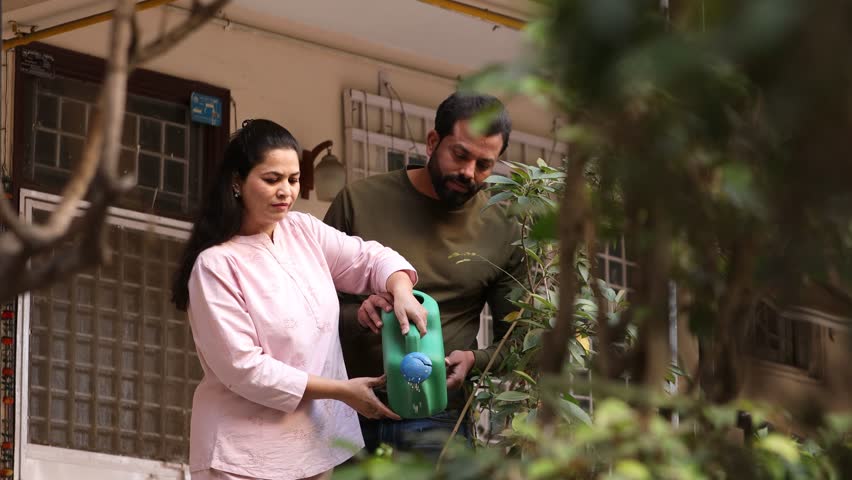 Young indian couple watering plants together outside their house garden in summer.Husband wife spend time together do Gardening hobby activity. Bonding and relationship. People of india. 4k