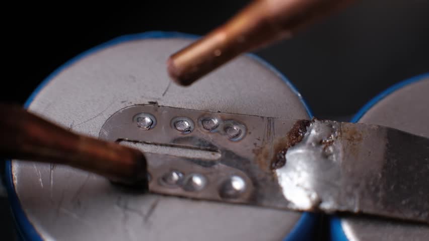 Macro slow motion footage shows copper electrodes creating sequential weld points on a battery cell tab, with bright sparks highlighting the resistance welding process.
