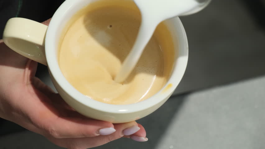 Barista professionally pours milk from mug into coffee in circular motion. Woman holds stream in particular place to create image on surface