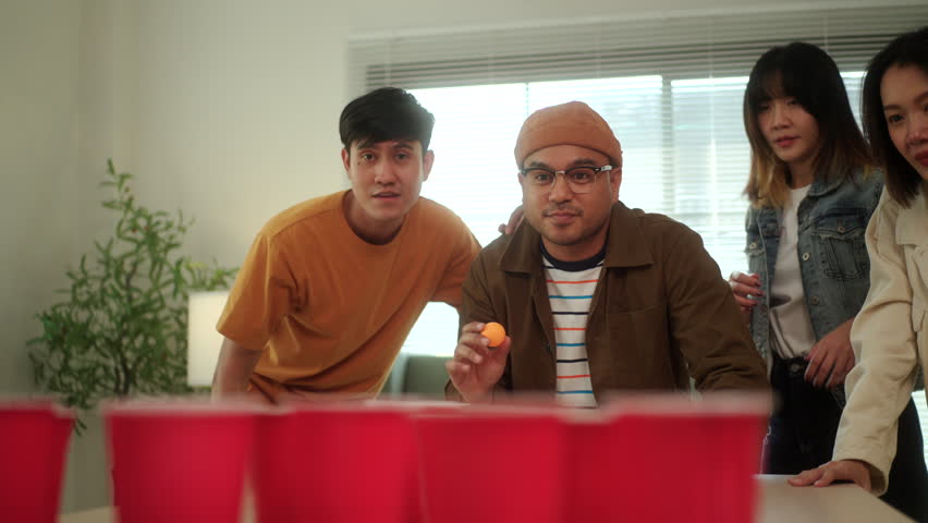 A group of young Asian friends is playing a fun game of ping pong ball toss into red cups at a lively party. The friends enjoy their time together, laughing and competing in the game.
