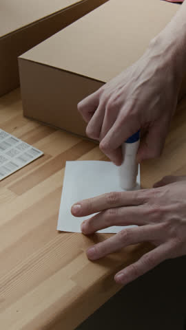 Vertical close-up view of hands of unrecognisable young man using glue stick to attach shipping label to cardboard box at workplace