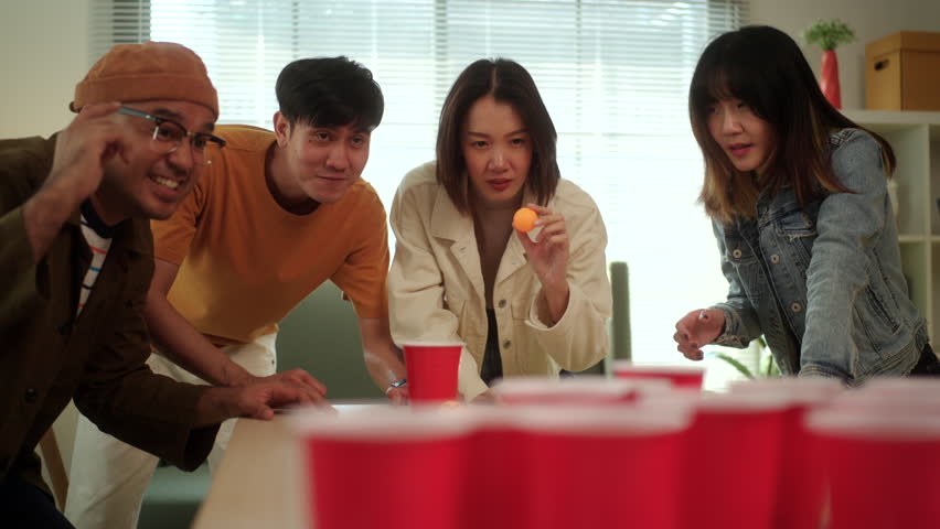 A group of young Asian friends is playing a fun game of ping pong ball toss into red cups at a lively party. The friends enjoy their time together, laughing and competing in the game.
