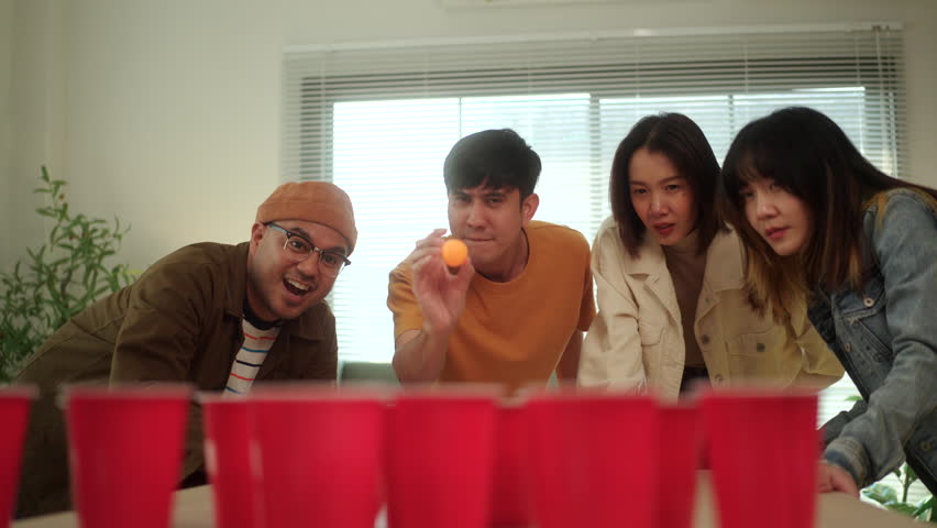 A group of young Asian friends is playing a fun game of ping pong ball toss into red cups at a lively party. The friends enjoy their time together, laughing and competing in the game.
