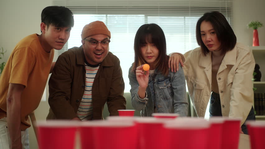A group of young Asian friends is playing a fun game of ping pong ball toss into red cups at a lively party. The friends enjoy their time together, laughing and competing in the game.

