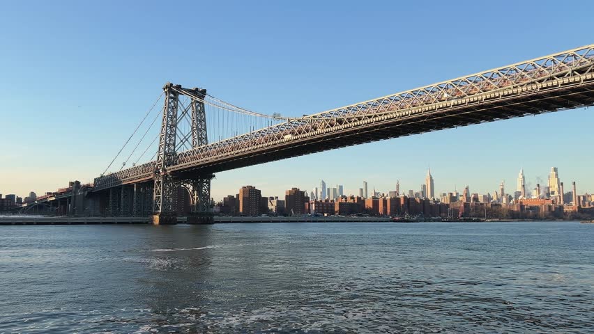 Sunny daytime point of view boat ride on East River, passing under Williamsburg Bridge. Yellow water taxi crosses, Manhattan skyline background. Travel, transport, establishing shot, USA
