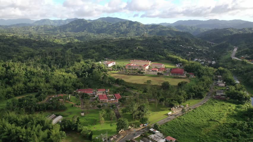 A drone landscape view of UPR - University of Puerto Rico in Utuado surrounded by green spaces and lush hills under cloudy sky