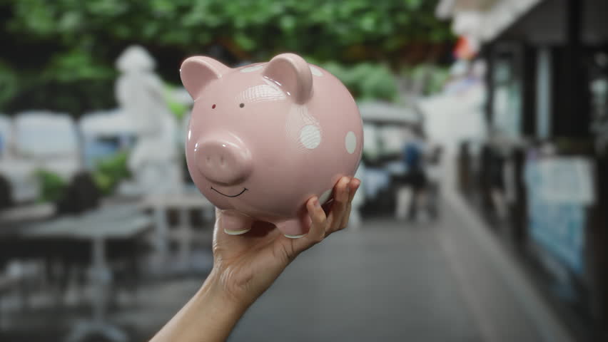 Hand holding pink piggy bank at outdoor city terrace against blurred urban background, symbolizing savings, finance, and investment in a bustling restaurant setting.