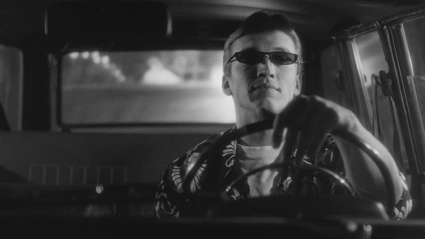 Young man wearing stylish sunglasses driving vintage car at night with blurred street lights in background. Retro cinematic noir atmosphere, black and white footage