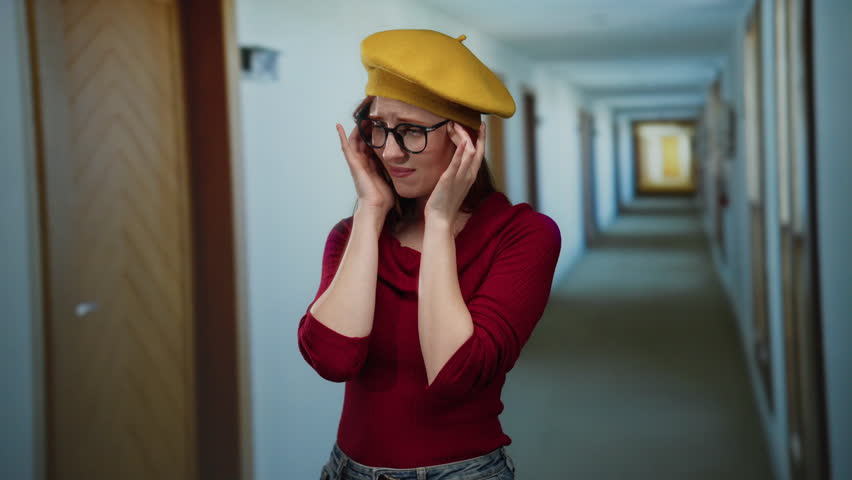 Woman with glasses and red hair stands thoughtfully in a hotel hallway, wearing a red sweater and yellow beret, capturing expressions of contemplation and style.