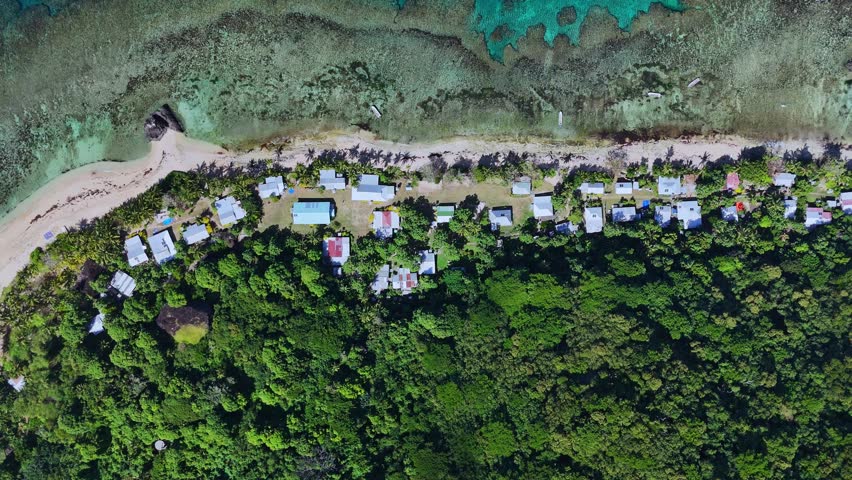 Aerial footage captures a traditional Fijian coastal village with small fishing boats anchored near the shore, surrounded by coral reefs and shallow turquoise waters.