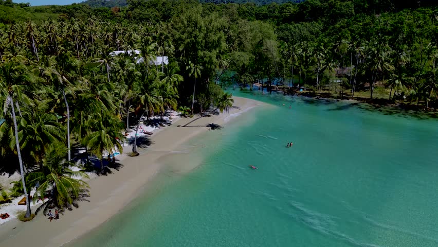 Explore a pristine beach in Koh Kood Thailand where turquoise waters meet white sands and swaying palm trees. Visitors enjoy swimming and relaxation under the sun in this tropical oasis.