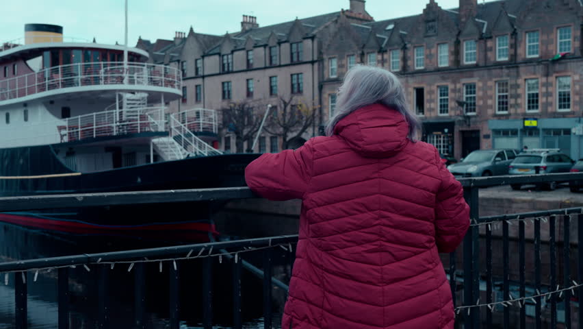 Grey-Haired Woman in Red Jacket Looking at Black Ship in Leith Port from Bridge Over River on Sunny Day in Edinburgh, Scotland