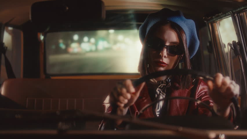 Stylish girl wearing blue beret, leather coat and sunglasses driving vintage car on street at night, with city lights in background