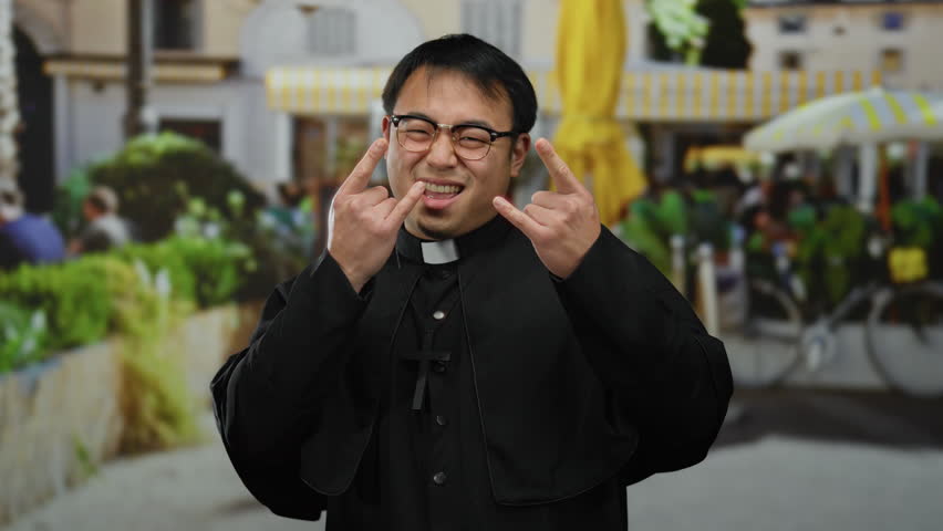 Asian man wearing priest outfit stands smiling in outdoor cafe with vibrant urban background, suggesting a joyful religious theme.