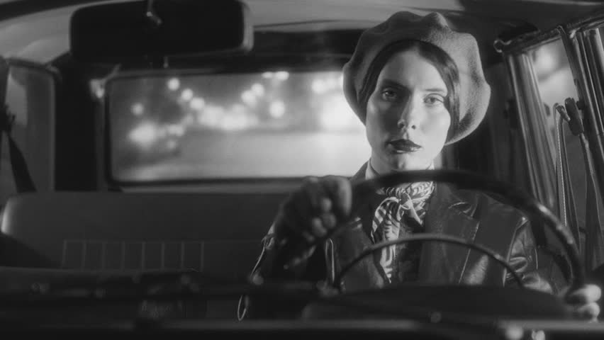 Beautiful woman in elegant retro outfit driving a vintage car on a city street at night. Black and white video portrait, classic film noir aesthetic