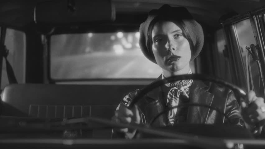 Beautiful woman in elegant retro outfit driving a vintage car on a city street at night. Black and white video portrait, classic film noir aesthetic