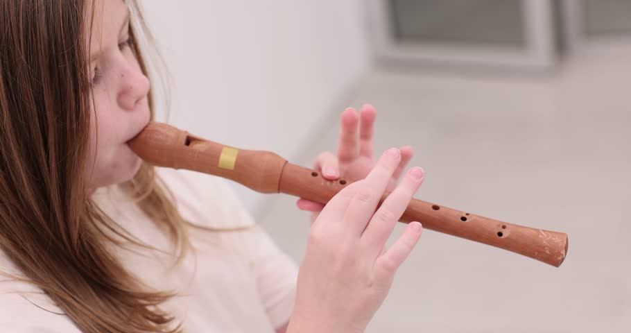 Focused schoolgirl with long hair plays wooden recorder in light studio room. Active preparation rehearsal of child before music performance on flute