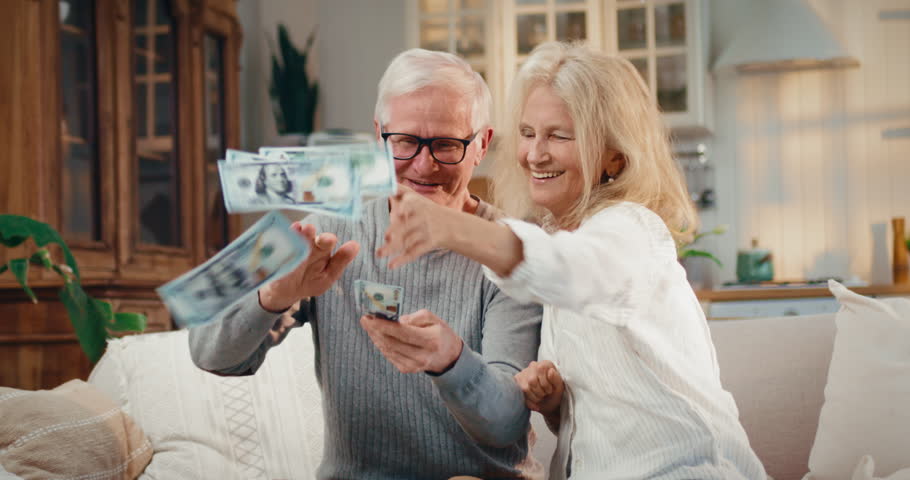 Happy old spouses throwing money around, portrait of rich people at home. Pension benefits for retirees, richness and wealth, aged woman and man playing with cash dollars, happy aging and retirement