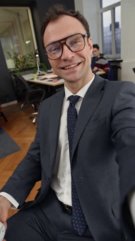 Vertical POV of male Caucasian gen Y employee in eyeglasses and formalwear talking to camera while vlogging at office