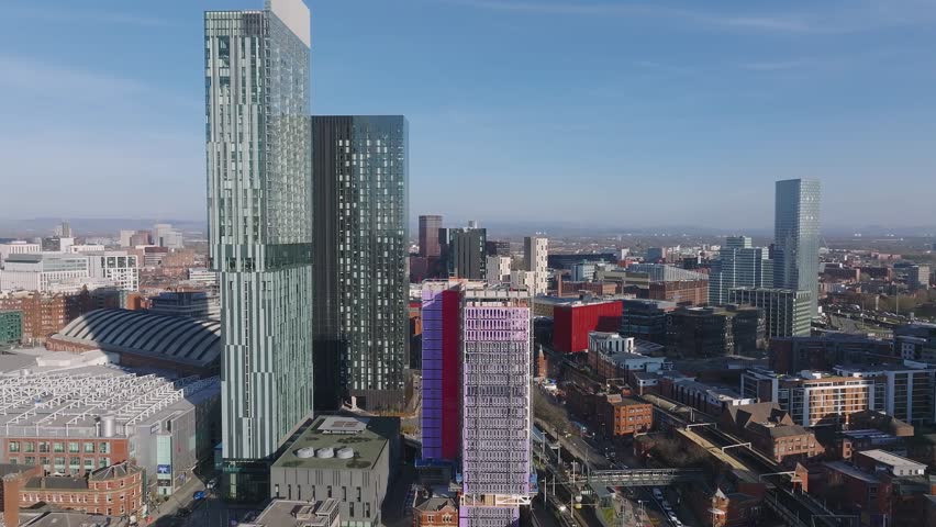 Aerial view of Manchester, UK, featuring skyscrapers like Beetham Tower, a red and white building under construction, train tracks, and a moving train.
