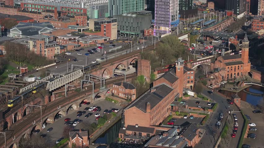 Aerial view of Manchester, UK, showing trains on tracks, canals, red brick buildings, modern high rises, and historic architecture in motion.