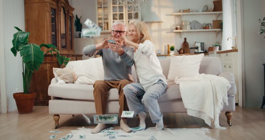 Financial success, happy old married couple throwing dollars banknotes at home. Rich people enjoying wealth, playing with cash money, grandfather and grandma spreading pension benefits, joyful smiles