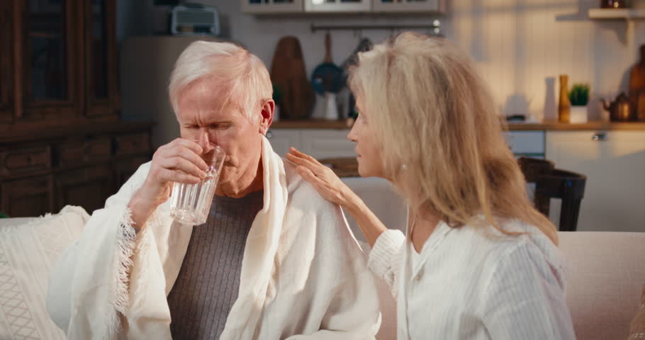 Old man feeling bad, woman giving him pills, healthcare and curing influenza. Medium portrait of two loving spouses in home interior, mature male person taking pill for preventing heart attack