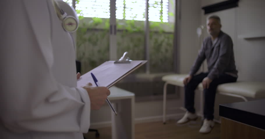 Female doctor holding clipboard with stethoscope as middle-aged male patient sits in background in clinic exam room