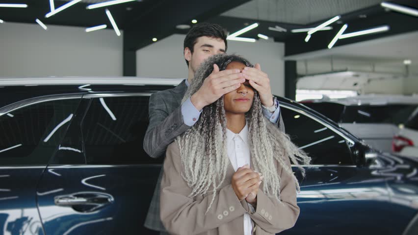 Man surprises woman with a car reveal in modern showroom filled with luxury vehicles during daylight hours