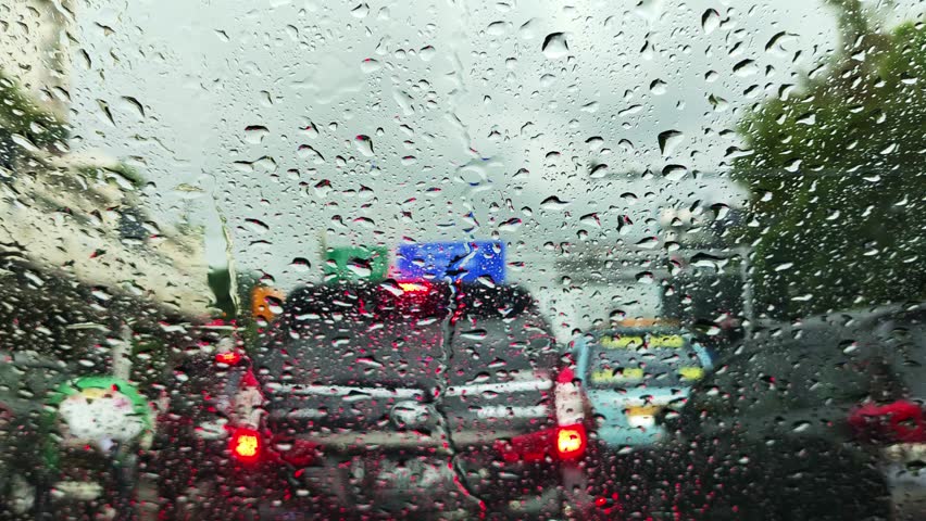 A car is driving down a wet road with raindrops on the windshield. The car is surrounded by other vehicles, including a truck and a motorcycle