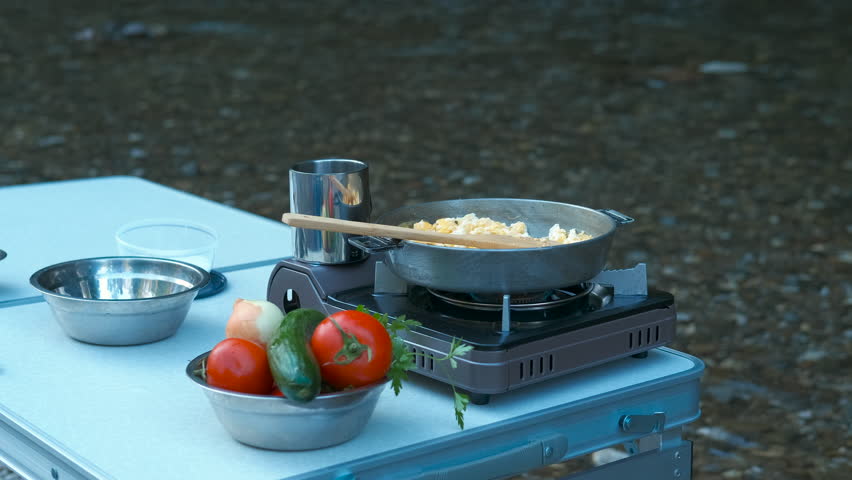 Cooking scrambled eggs at a campsite. Preparing scrambled eggs on portable gas stove while surrounded by fresh vegetables at rustic outdoor campsite, showcasing wilderness cooking experience