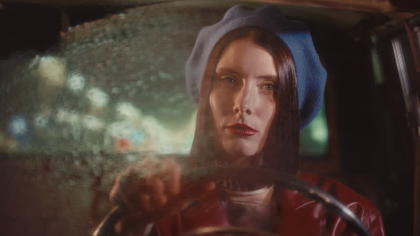Attractive girl in retro-style outfit driving vintage car on rainy night with raindrops on windshield and soft bokeh lights in background