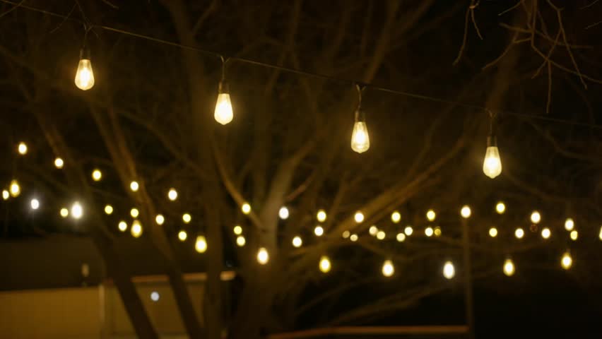 Warm glowing string lights hanging across bare tree branches on a terrace at night. Cozy outdoor evening atmosphere, perfect for celebrations, weddings, or romantic gatherings. 4k footage