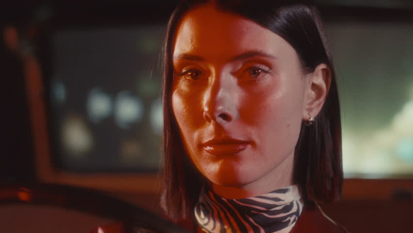 Young pretty woman with red lipstick and patterned scarf driving vintage car at night, looking thoughtfully at road. Close-up shot
