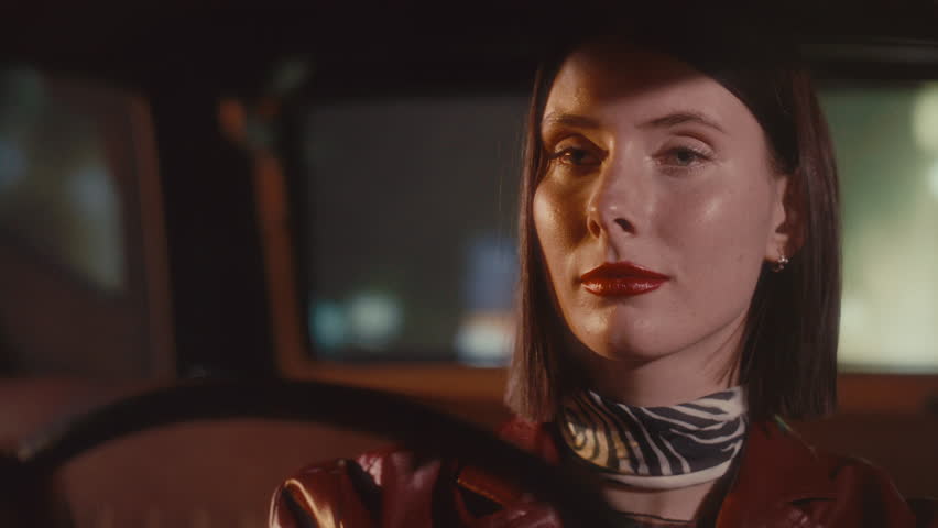 Young feminine woman with red lips driving classic retro car on city street and looking at night road