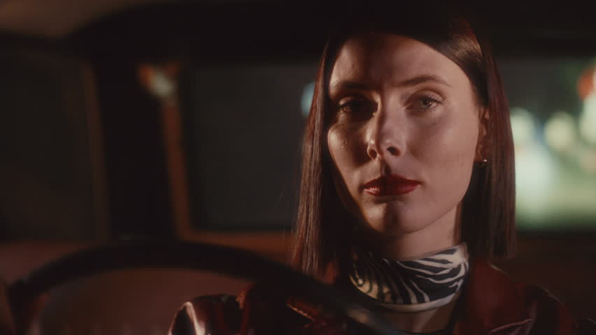 Young beautiful woman with red lips looking confidently into camera while driving retro car through night. Video portrait