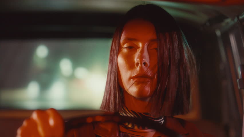 Young attractive woman looking into camera, with hair blowing across her face as she driving vintage car through city at night. Video portrait